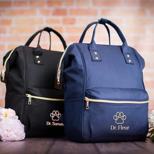 May include: Two personalized pet backpacks. One is black with gold zippers and the other is navy blue with gold zippers. Each backpack has a paw print and a name embroidered on the front. The backpacks have top handles and adjustable straps.