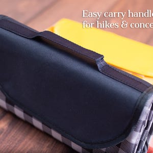 Puede incluir: Manta de picnic plegada con asa y solapa azul marino, con un estampado de cuadros blancos y negros. La manta es parcialmente visible, con una manta a cuadros amarilla y naranja en el fondo. Texto: Easy carry handle, for hikes & concerts.