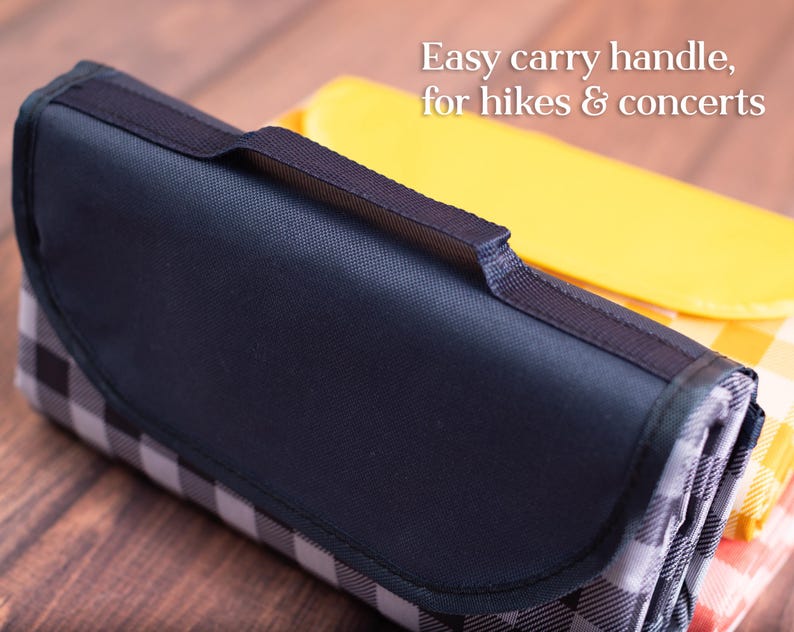 Puede incluir: Manta de picnic plegada con asa y solapa azul marino, con un estampado de cuadros blancos y negros. La manta es parcialmente visible, con una manta a cuadros amarilla y naranja en el fondo. Texto: Easy carry handle, for hikes & concerts.