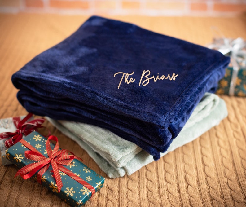 May include: A navy blue fleece blanket with the text "The Briars" embroidered in gold. The blanket is folded and placed on a brown cable knit surface.