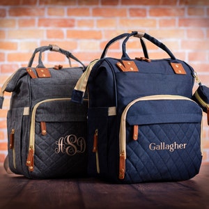 May include: Two structured backpacks with top handles and quilted front pockets. The charcoal gray bag features the monogram ASD, while the navy blue bag displays the name Gallagher. Both include tan leather-like accents, gold zippers, and side pockets.