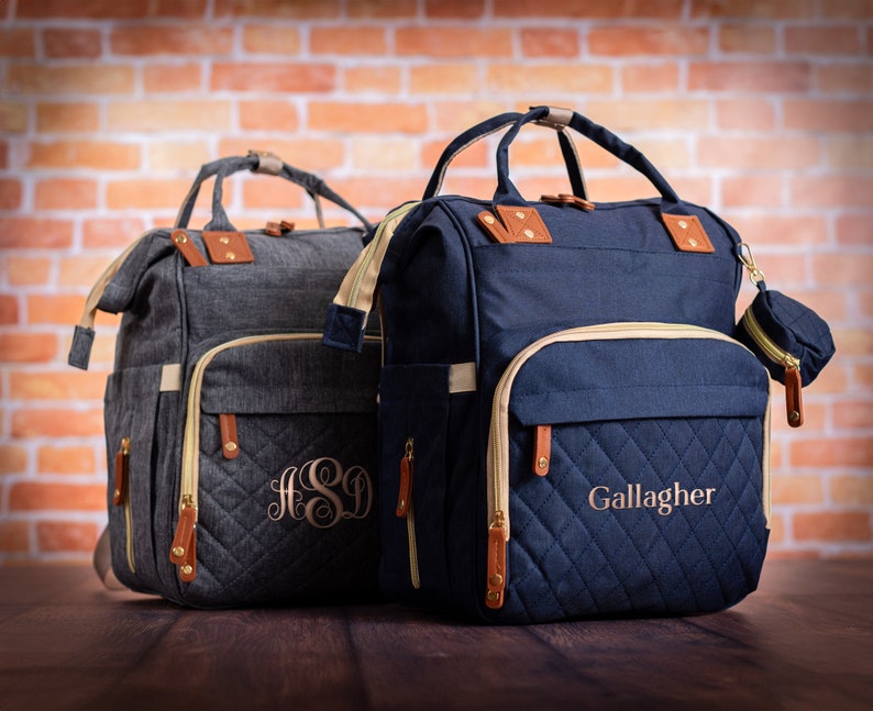 May include: Two structured backpacks with top handles and quilted front pockets. The charcoal gray bag features the monogram ASD, while the navy blue bag displays the name Gallagher. Both include tan leather-like accents, gold zippers, and side pockets.