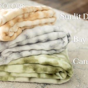 May include: A stack of three plush blankets in the "Watercolors" collection. The top blanket is beige and tan, the middle is gray and white, and the bottom is green and white. The blankets are labeled "Sunlit Dune", "Bay Fog", and "Canopy Swirl".