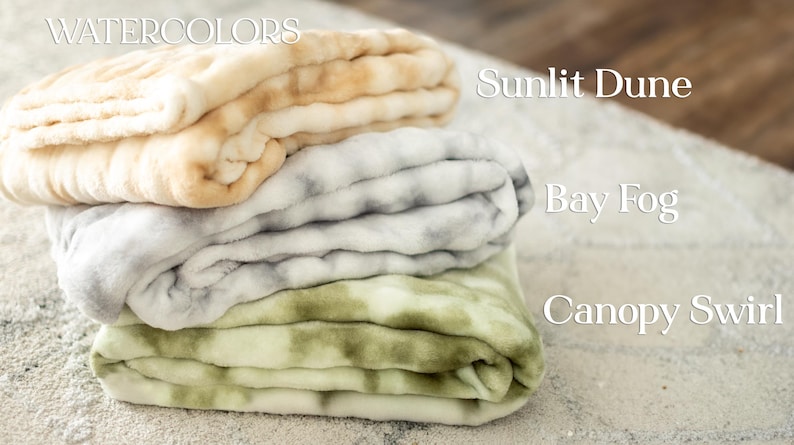 May include: A stack of three plush blankets in the "Watercolors" collection. The top blanket is beige and tan, the middle is gray and white, and the bottom is green and white. The blankets are labeled "Sunlit Dune", "Bay Fog", and "Canopy Swirl".