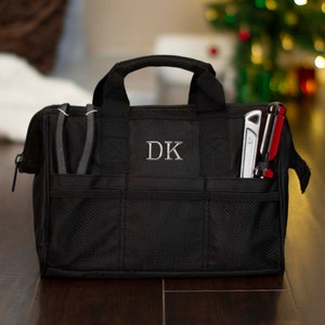 K&ouml;nnte beinhalten: Schwarze Werkzeugtasche mit Rei&szlig;verschluss und mehreren Taschen. Die Tasche hat eine Netztasche und ein Monogramm "DK" auf der Vorderseite.