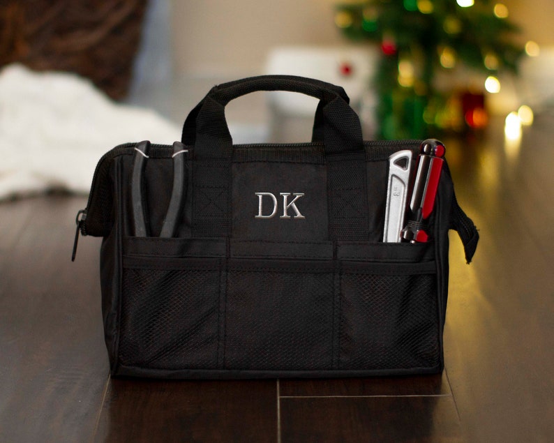 K&ouml;nnte beinhalten: Schwarze Werkzeugtasche mit Rei&szlig;verschluss und mehreren Taschen. Die Tasche hat eine Netztasche und ein Monogramm "DK" auf der Vorderseite.