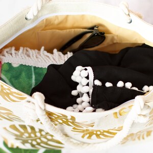 May include: A white canvas tote bag with gold tropical leaf print. The bag is open and filled with items, including a black shirt with white pom pom trim, a green and pink beach towel, and sunglasses.