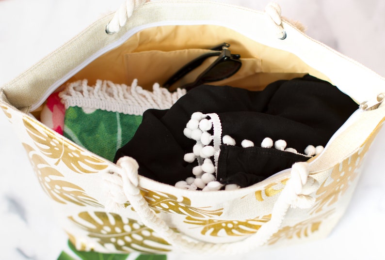 May include: A white canvas tote bag with gold tropical leaf print. The bag is open and filled with items, including a black shirt with white pom pom trim, a green and pink beach towel, and sunglasses.
