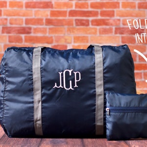 May include: A navy blue travel duffel bag with gray straps and the monogram "JCP" embroidered on the front. The bag folds into a small, compact pouch.