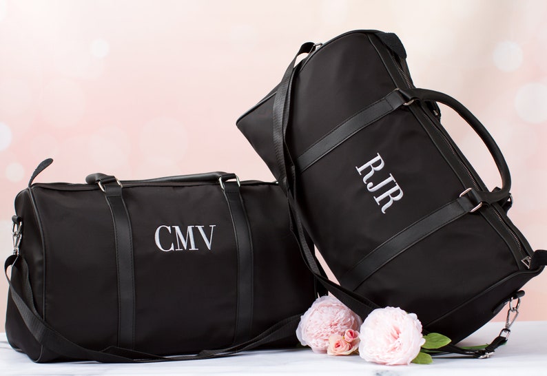 以下が含まれることがあります： 白い刺繍で「CMV」と「RJR」のイニシャルがパーソナライズされた、革のアクセントが付いた黒いダッフルバッグ2つ。バッグはピンクの花が咲いた白い表面の上に置かれています。
