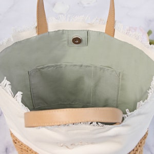 May include: A white canvas tote bag with a frayed edge and a brown leather handle. The bag has a green fabric lining with a small pocket.