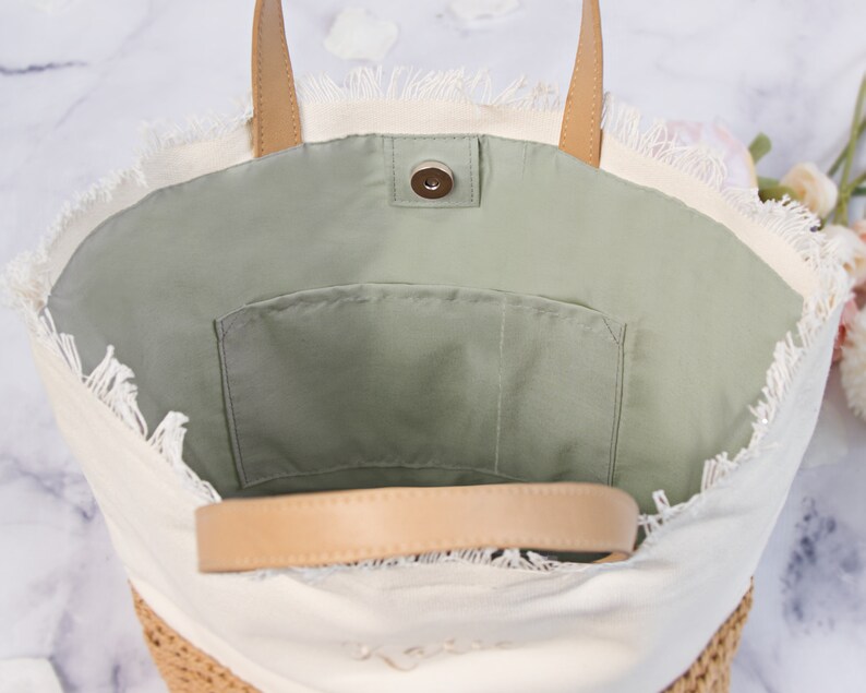 May include: A white canvas tote bag with a frayed edge and a brown leather handle. The bag has a green fabric lining with a small pocket.