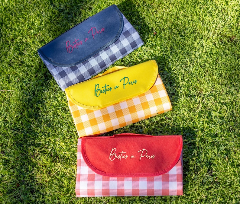 Puede incluir: Tres mantas de picnic con estampados de cuadros en azul marino, amarillo y rojo. Cada manta tiene una solapa a juego con el texto "Besties in Paris" en cursiva. Las mantas est&aacute;n dobladas y colocadas sobre hierba verde.