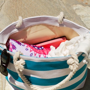 Puede incluir: Una bolsa de playa a rayas con rayas blancas y azul turquesa y asas de cuerda. La bolsa est&aacute; abierta y llena de art&iacute;culos, incluyendo una toalla estampada rosa y blanca, una toalla blanca y un libro rosa.