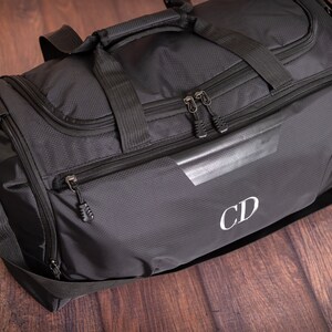 K&ouml;nnte beinhalten: Schwarze Reisetasche mit einer Rei&szlig;verschlusstasche und einem wei&szlig;en Monogramm "CD" auf der Vorderseite.
