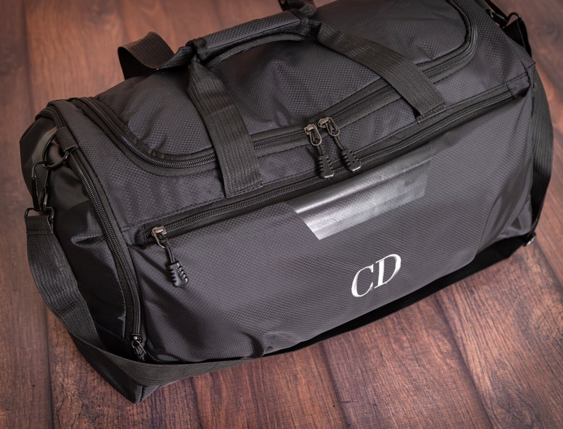 K&ouml;nnte beinhalten: Schwarze Reisetasche mit einer Rei&szlig;verschlusstasche und einem wei&szlig;en Monogramm "CD" auf der Vorderseite.