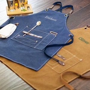 May include: Two personalized denim aprons with adjustable straps. The blue apron has a white embroidered name "Erickson" and the brown apron has a green embroidered name "Oliver".