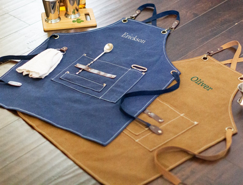 May include: Two personalized denim aprons with adjustable straps. The blue apron has a white embroidered name "Erickson" and the brown apron has a green embroidered name "Oliver".