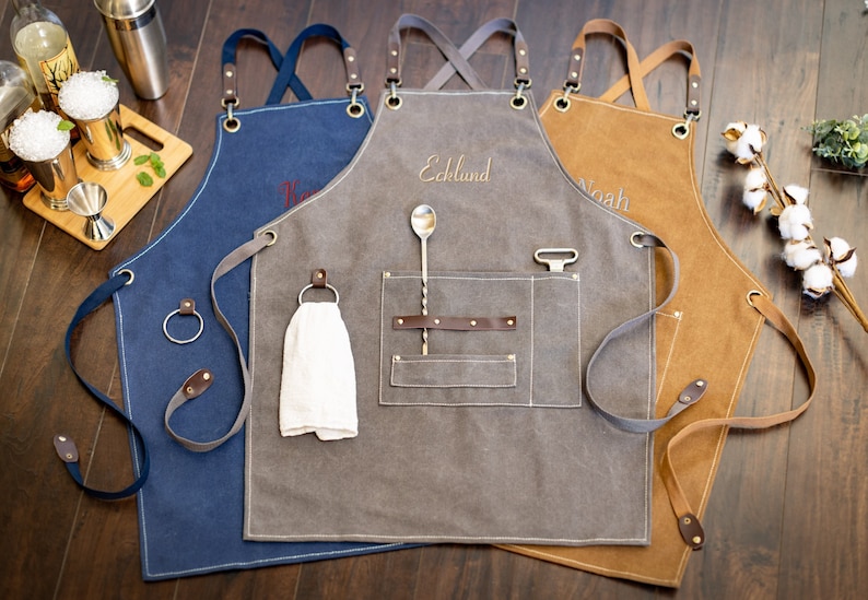 May include: Three personalized canvas aprons with adjustable straps and leather accents. The aprons are in blue, gray, and brown. The gray apron has a pocket with a bar spoon and a cocktail shaker. The aprons are embroidered with the names "Noah", "Ecklund", and "K", respectively.