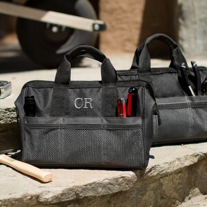 Puede incluir: Dos bolsas de herramientas de lona negra con m&uacute;ltiples bolsillos y un compartimento de malla. La bolsa de la izquierda tiene las iniciales "CR" bordadas en la parte delantera.