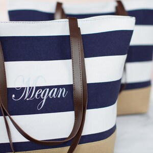 Puede incluir: Una bolsa de lona a rayas blancas y azul marino con asas de cuero marr&oacute;n. La bolsa tiene un nombre bordado personalizado, "Megan", en hilo blanco.