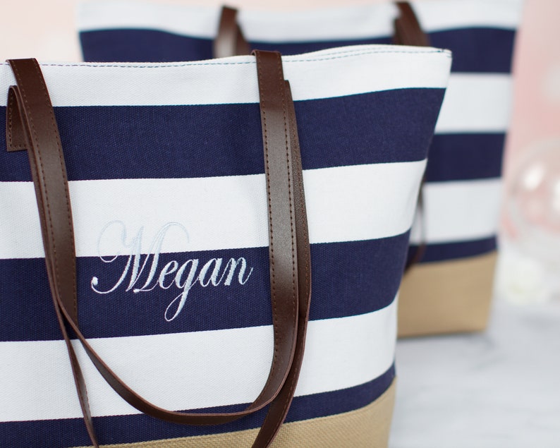 Puede incluir: Una bolsa de lona a rayas blancas y azul marino con asas de cuero marr&oacute;n. La bolsa tiene un nombre bordado personalizado, "Megan", en hilo blanco.