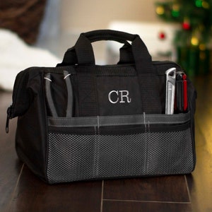 K&ouml;nnte beinhalten: Schwarze Werkzeugtasche aus Canvas mit Rei&szlig;verschluss, Netzfach und grauem Besatz. Die Tasche hat einen Griff und ist mit den Initialen "CR" bestickt.
