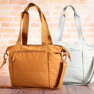 May include: Two tote bags. One is a mustard yellow color, the other is a light gray. Both bags have top handles and adjustable shoulder straps. The bags have a zippered top closure and a front zippered pocket. The background is a brick wall.