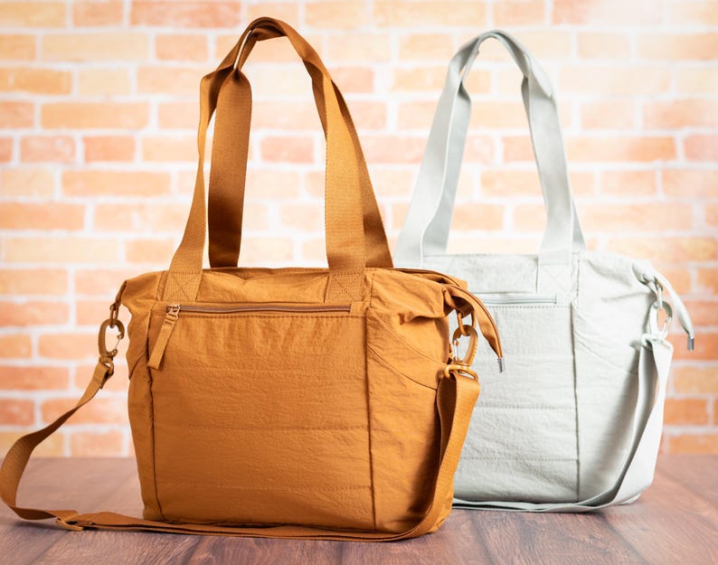 May include: Two tote bags. One is a mustard yellow color, the other is a light gray. Both bags have top handles and adjustable shoulder straps. The bags have a zippered top closure and a front zippered pocket. The background is a brick wall.