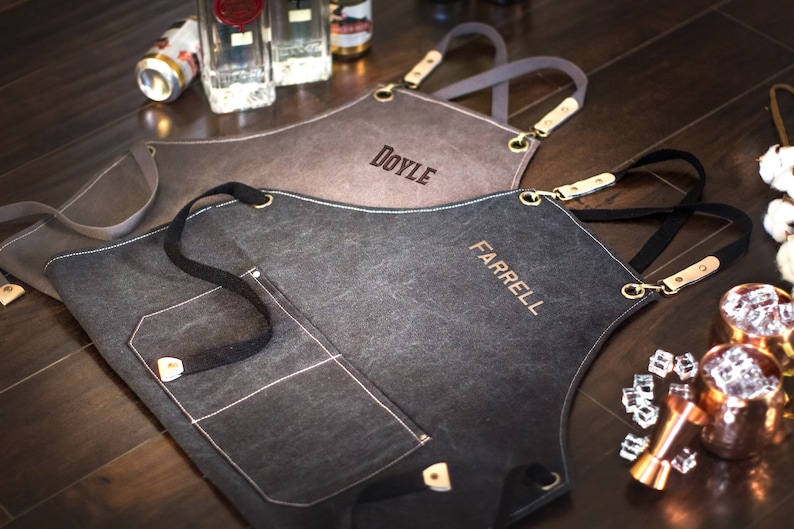 May include: Two personalized black canvas aprons with adjustable straps and a large pocket. The aprons are embroidered with the names "DOYLE" and "FARRELL".