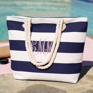 Puede incluir: Una bolsa de lona de rayas blancas y azul marino con asas de cuerda. La bolsa tiene un monograma rosa "CMW" bordado en la parte delantera.