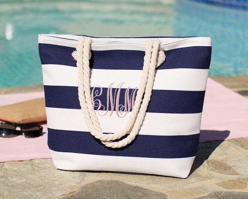 Puede incluir: Una bolsa de lona de rayas blancas y azul marino con asas de cuerda. La bolsa tiene un monograma rosa "CMW" bordado en la parte delantera.