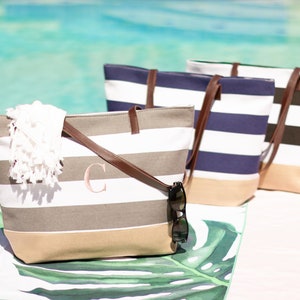 Puede incluir: Tres bolsas de lona a rayas con asas de cuero. Las bolsas est&aacute;n alineadas en una fila sobre una superficie con estampado blanco y verde. La bolsa de la izquierda tiene un patr&oacute;n de rayas beige y blanco y un monograma con la letra rosa "C" en la parte delantera. La bolsa del medio tiene un patr&oacute;n de rayas azul marino y blanco. La bolsa de la derecha tiene un patr&oacute;n de rayas negras y blancas.