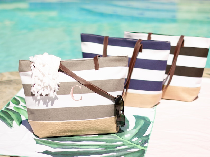 Puede incluir: Tres bolsas de lona a rayas con asas de cuero. Las bolsas est&aacute;n alineadas en una fila sobre una superficie con estampado blanco y verde. La bolsa de la izquierda tiene un patr&oacute;n de rayas beige y blanco y un monograma con la letra rosa "C" en la parte delantera. La bolsa del medio tiene un patr&oacute;n de rayas azul marino y blanco. La bolsa de la derecha tiene un patr&oacute;n de rayas negras y blancas.