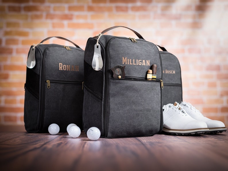 May include: Three black canvas golf shoe bags with personalised names, "Ronan", "Milligan", and "Bosch". The bags have a zipped pocket and a shoe horn attached. Three white golf balls are in front of the bags. A pair of white golf shoes are in front of the bag with the name "Bosch".