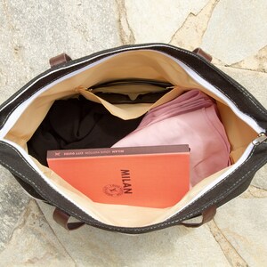 May include: A black canvas tote bag with a white zipper and brown leather handles. The bag is open and filled with items, including a book titled "MILAN LOUIS VUITTON CITY GUIDE", sunglasses, and a pink scarf.