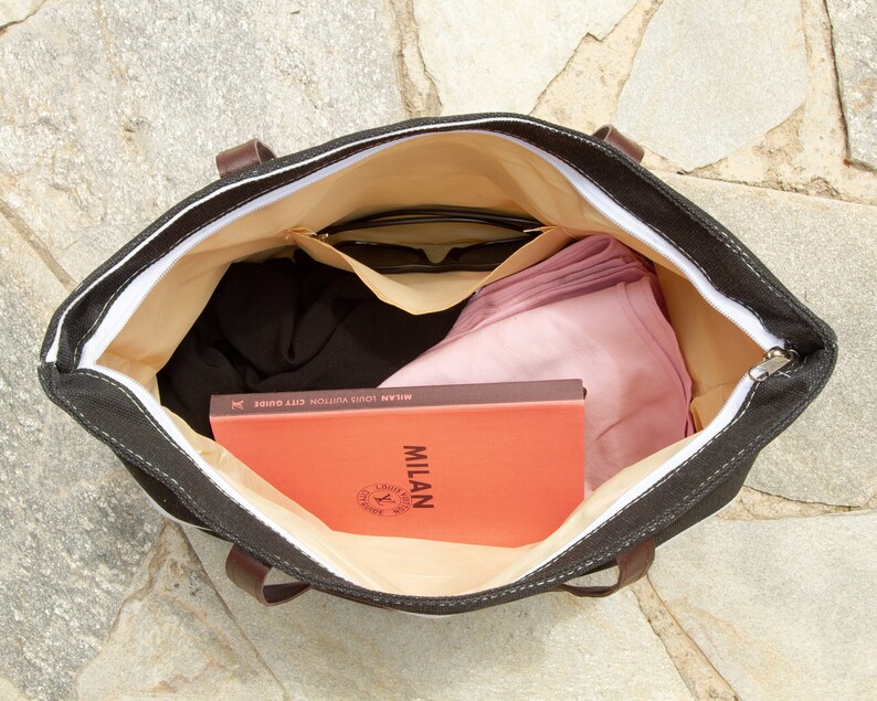 May include: A black canvas tote bag with a white zipper and brown leather handles. The bag is open and filled with items, including a book titled "MILAN LOUIS VUITTON CITY GUIDE", sunglasses, and a pink scarf.