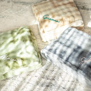 May include: Three personalized faux fur blankets in shades of green, beige, and gray. Each blanket is folded and embroidered with a name in a contrasting color: "Gianna" in teal, "Margot" in white, and "Sydney" in pink. The blankets have a soft, plush texture.