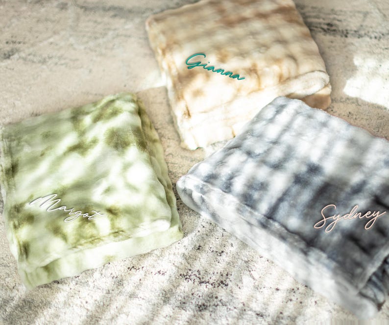 May include: Three personalized faux fur blankets in shades of green, beige, and gray. Each blanket is folded and embroidered with a name in a contrasting color: "Gianna" in teal, "Margot" in white, and "Sydney" in pink. The blankets have a soft, plush texture.