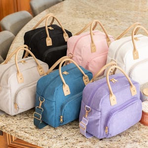 May include: A collection of six corduroy travel bags in various colors: black, pink, white, gray, teal, and purple. Each bag has a top handle, a detachable shoulder strap, and a front pocket. The handles and accents are a light tan color.