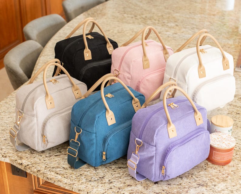 May include: A collection of six corduroy travel bags in various colors: black, pink, white, gray, teal, and purple. Each bag has a top handle, a detachable shoulder strap, and a front pocket. The handles and accents are a light tan color.