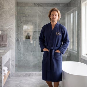May include: A navy blue waffle-knit robe featuring long sleeves, a tie waist, two front pockets, and the embroidered monogram letters ASL in a light tan thread on the left chest area. The garment has a shawl collar and a textured fabric finish.