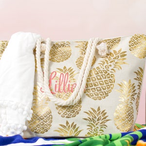 May include: A white canvas tote bag with gold pineapple print and rope handles. The bag is personalized with the name "Lillie" in pink lettering. A white towel with pom-poms is in the bag.