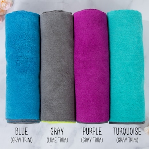 May include: Four rolled up towels in different colors: blue with gray trim, gray with lime trim, purple with gray trim, and turquoise with gray trim. The towels are arranged in a row on a white surface.