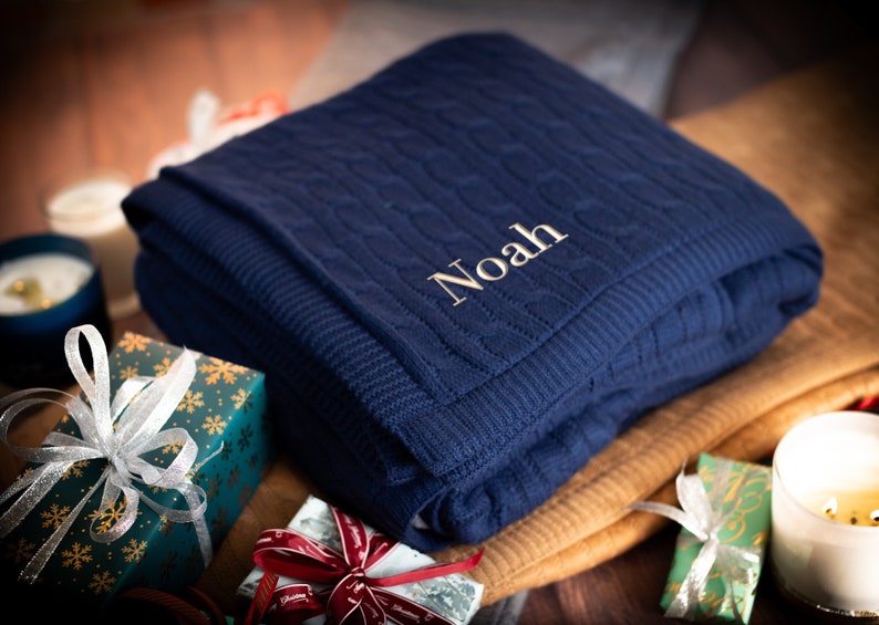 Pode incluir: Uma manta de malha azul marinho com cabo e o nome "Noah" bordado em branco na frente. A manta est&aacute; dobrada e colocada sobre uma superf&iacute;cie marrom com presentes embrulhados e velas.