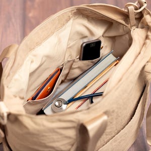 May include: Beige tote bag with multiple pockets, containing a smartphone, a book, a stethoscope, and a wallet. The bag has a shoulder strap and is made of a light-colored fabric. The interior is lined with a lighter shade of beige.