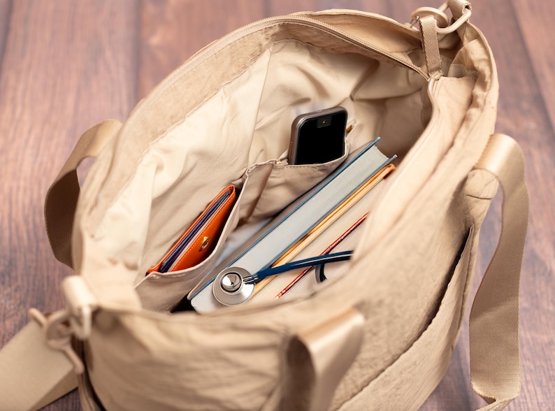 May include: Beige tote bag with multiple pockets, containing a smartphone, a book, a stethoscope, and a wallet. The bag has a shoulder strap and is made of a light-colored fabric. The interior is lined with a lighter shade of beige.