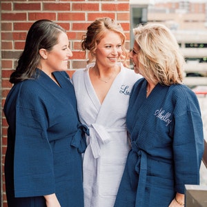May include: Three waffle-knit robes featuring long sleeves and tie-waist closures. Two robes are dark navy blue, and one robe is white. The white robe features the embroidered name "Linda" and the navy robe features the embroidered name "Shelley".