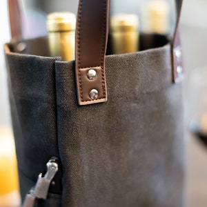 May include: A black canvas wine tote bag with brown leather handles and metal rivets. The bag is filled with wine bottles.