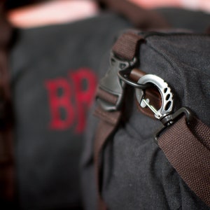 K&ouml;nnte beinhalten: Eine schwarze Canvas-Reisetasche mit braunen Riemen und einem silbernen Metallhaken. Die Tasche hat ein rotes gesticktes Logo mit der Aufschrift "BR".